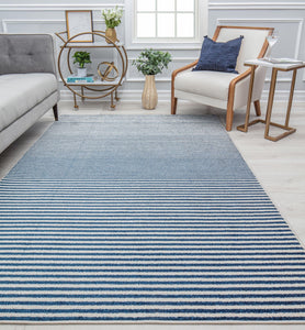 Contemporary striped rug in shades of blue and white, placed in a cozy living room with a gray sofa, wooden chair, and decor accents.