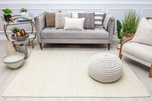 Cozy living room setup with a beige woven rug, gray sofa, decorative pillows, and a knitted pouf, showcasing a warm and inviting atmosphere.