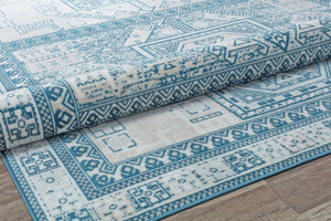 Close-up of a folded beige and blue geometric patterned rug on wooden floor, showcasing intricate design and texture.