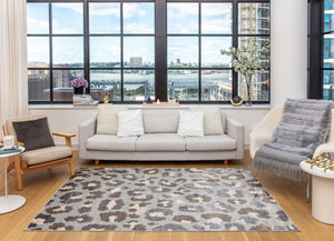 Bright living room with large windows, neutral-colored furniture, gray and white patterned rug, and city view in the background.