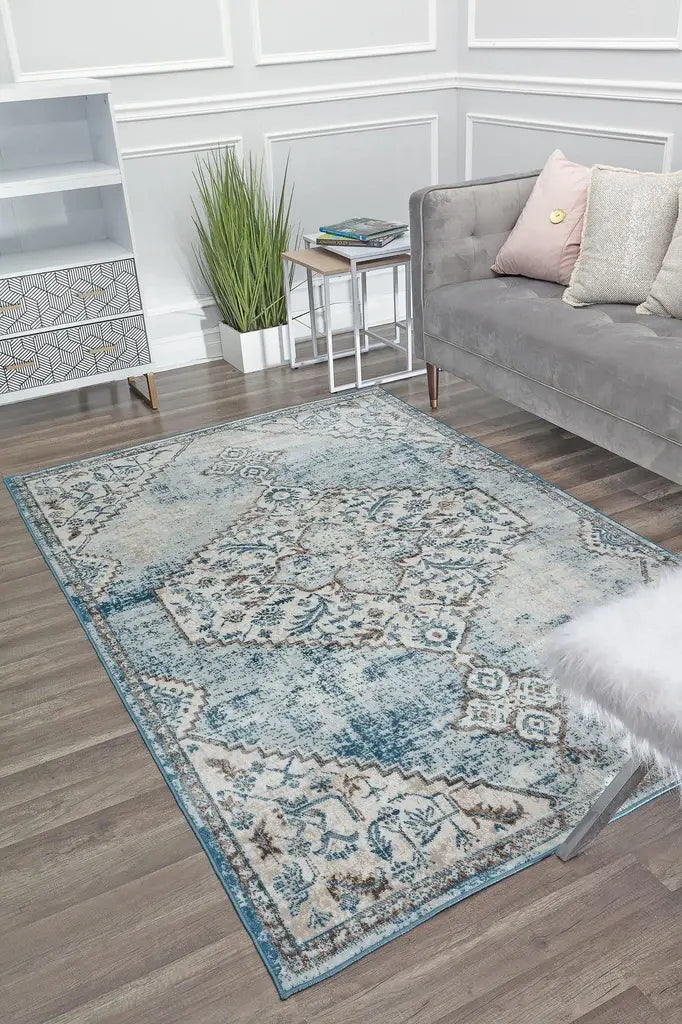 Elegant living room with a gray sofa, blue and white patterned rug, wooden floor, green plant, side tables, and a small bookcase