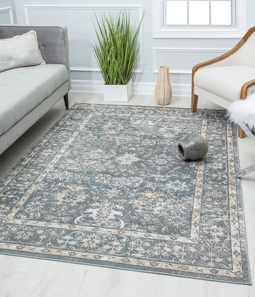 Living room featuring a vintage-style dark floral rug, a gray sofa, white armchair, green plant, and decorative vase against white walls