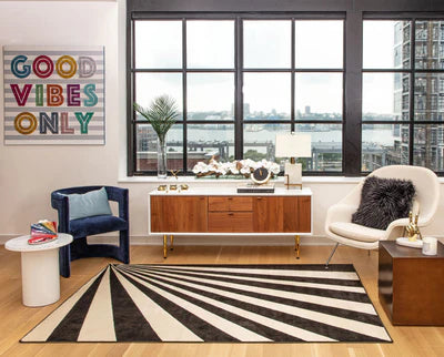 Modern living room with a black and white striped rug, a blue armchair, white chair, wooden console, and a 'Good Vibes Only' wall art.