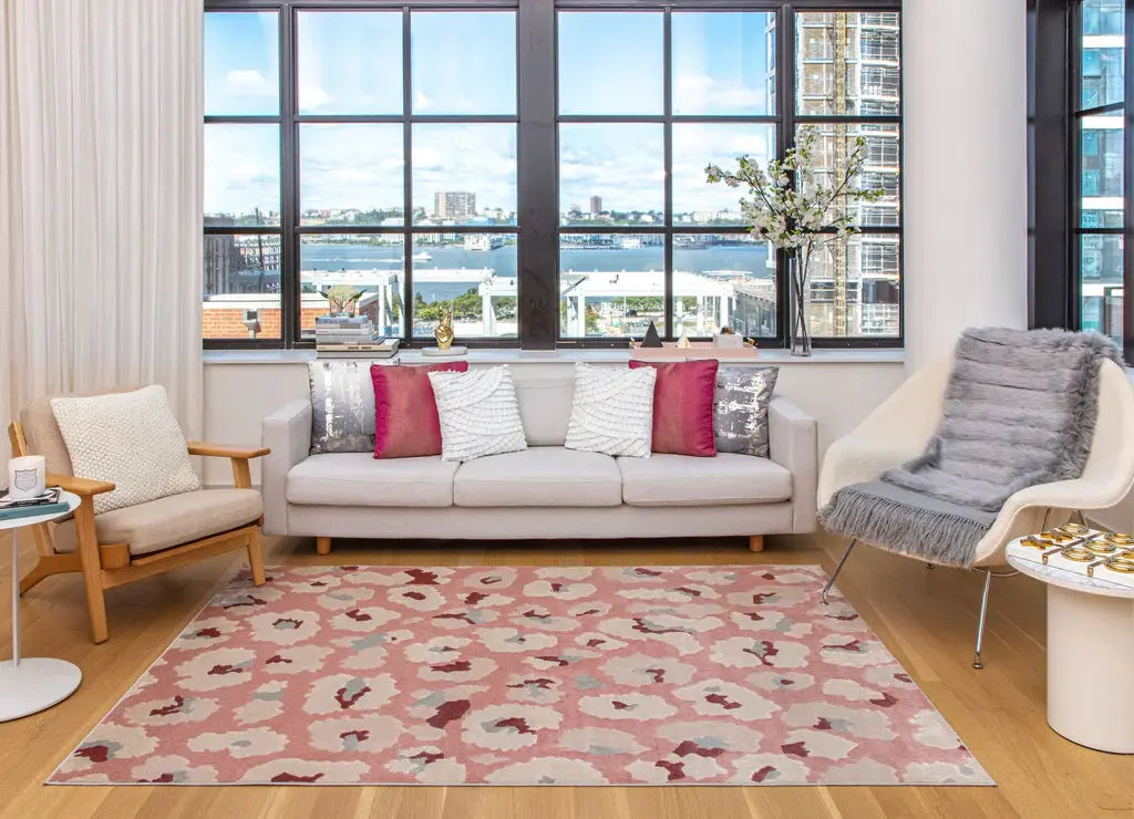 Modern living room with large windows, stylish white furniture, pink abstract rug, and vibrant cushions overlooking a cityscape.